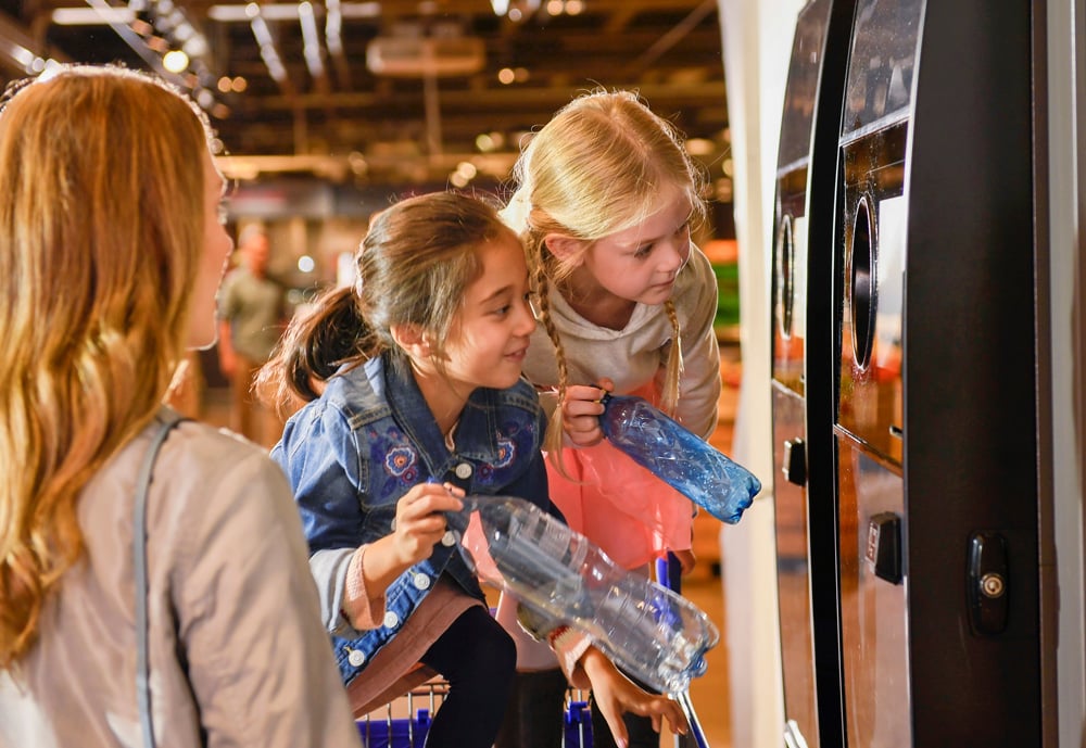 image of two girls putting bottles in a reverse vending machine while their mother is watching them