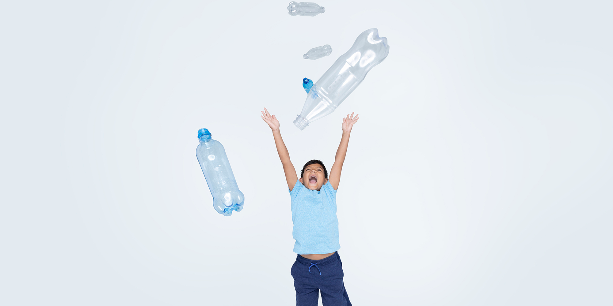 Young boy in light blue shirt and dark blue shorts with big smile and arms outstretched above his head and several empty plastic beverage containers floating in the air around him. 