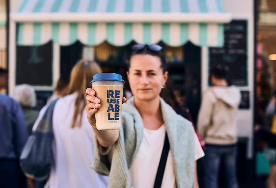 Woman in t-shirt holding a coffee drink in a clear REUSEABLE cup in the street outside a coffee shop