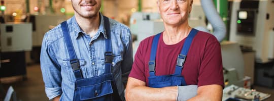 Porträt zweier Mechaniker in Overalls, die an ihrem Arbeitsplatz an der Produktionsanlage stehen, mit einer Drehbank im Hintergrund