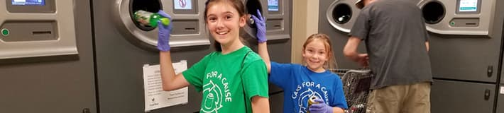 Two smiling young girls wearing gloves while using reverse vending machines